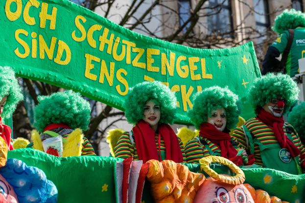 Eine Gruppe von Clowns in bunten Kostümen marschiert durch eine Straße, hält ein Banner mit Text, mit Bäumen und einem Gebäude im Hintergrund.
