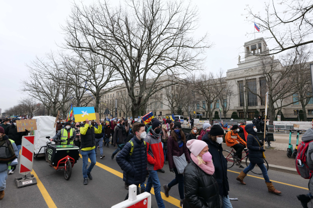 Eine große Gruppe von Menschen marschiert auf einer Straße bei einer Demonstration, einige halten Schilder und andere fahren Fahrräder, mit Bäumen und einem Gebäude im Hintergrund bei einem klaren blauen Himmel.