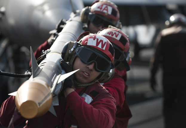 Eine Gruppe von Männern in roten Uniformen und Helmen steht zusammen, einer hält eine Rakete, im Hintergrund ist ein Flugzeug zu sehen.