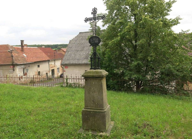 Ein hölzernes Kreuzdenkmal steht in einem von einem Metallzaun umgebenen Grasfeld, mit Bäumen, Häusern und einem bewölkten Himmel im Hintergrund.
