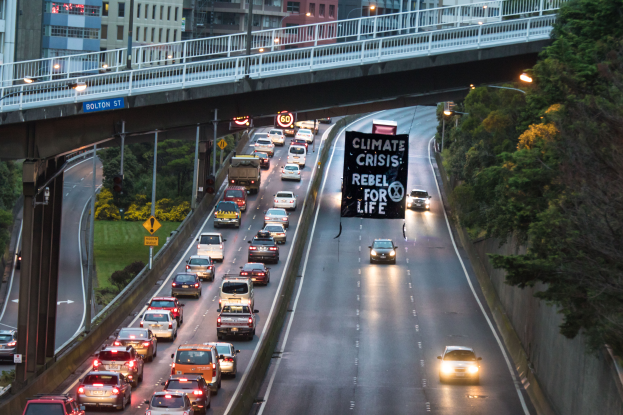 Eine belebte Stadtstraße mit starkem Verkehr, eine Brücke im Hintergrund und ein Banner mit Text in der Mitte der Straße, flankiert von Bäumen, Polen, Laternen, Schildern und Gebäuden auf der rechten Seite.