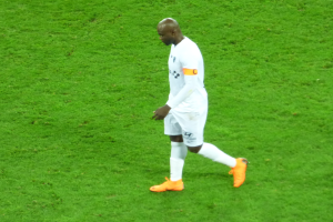 Ein Fußballspieler in einem weißen T-Shirt, weißen Shorts und orangefarbenen Schuhen, der über ein saftig grünes Feld geht.