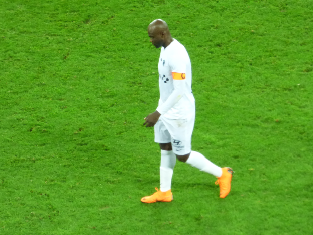 Ein Fußballspieler in einem weißen T-Shirt, weißen Shorts und orangefarbenen Schuhen, der über ein saftig grünes Feld geht.