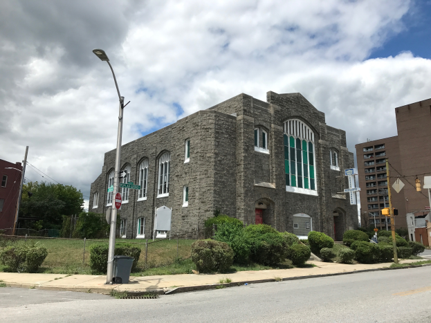 Eine alte Kirche an einer Straßecke, umgeben von Gebäuden, Straßeleitpfosten, Straßeleuchten, Schildern, Verkehrspfosten, Verkehrssignalen, Kraftfahrzeugen, Bühsen, Bäumen und einem bewölktem Himmel.
