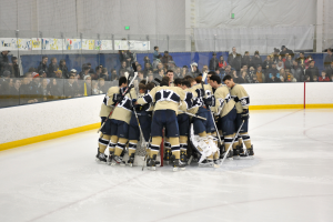 Eine Gruppe von Hockey-Spielern in Uniformen steht zusammen auf dem Eis mit Hockey-Schlägern, während Zuschauer von den Tribünen dahinter zusehen und ein Netz an der Wand sichtbar ist.