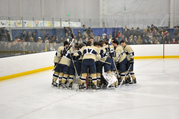 Eine Gruppe von Hockey-Spielern in Uniformen steht zusammen auf dem Eis mit Hockey-Schlägern, während Zuschauer von den Tribünen dahinter zusehen und ein Netz an der Wand sichtbar ist.