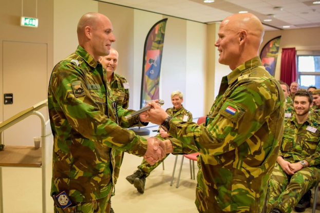 Zwei Männer in Militäruniformen geben sich die Hand vor einer sitzenden Gruppe, einem Tisch, einem Geländer, Bannern, einer Tür, einem Schild, einem Fenster mit Vorhang und Deckenleuchten im Hintergrund.