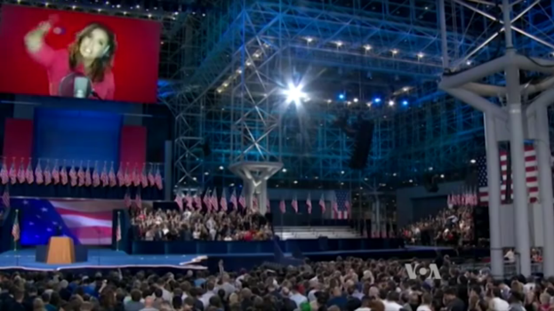 Eine große Menschenmenge sitzt vor einer Bühne mit einem Rednerpult, Mikrofon, Flaggen, einem Bildschirm mit dem Bild einer Frau und Beleuchtungsausrüstung, wahrscheinlich bei einer Präsidentschaftsdebatte.