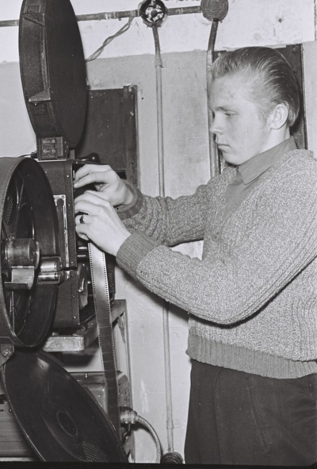 Ein Mann in weißem Hemd und dunkler Hose steht ernst vor einem alten schwarz-weißen Filmprojektor vor einer einfarbigen Wand.