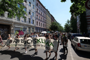Eine Gruppe von Menschen marschiert auf einer Straße und hält ein Banner hoch, mit parkenden Autos auf der rechten Seite, einem Radfahrer auf der linken Seite und Gebäuden, Bäumen und einem Schild im Hintergrund unter einem sichtbaren Himmel.
