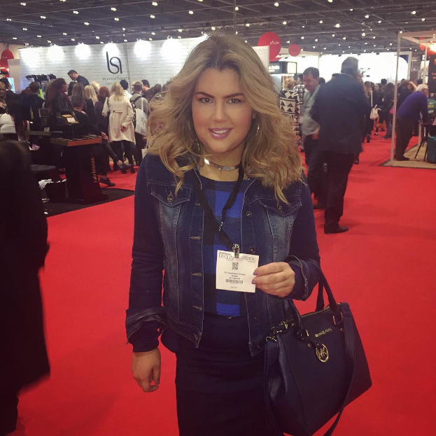 Eine Frau steht auf einem roten Teppich, lächelt und hält eine Handtasche, umgeben von Menschen, Lichtern und Schildern auf einer Modenschau.