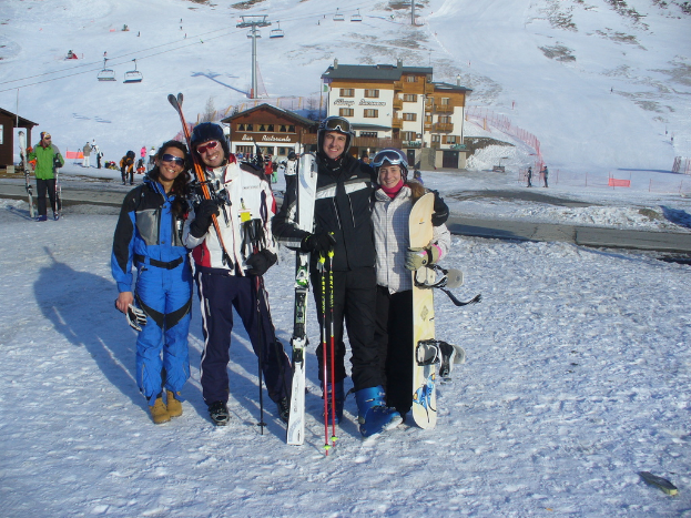 Vier Personen posieren für ein Foto mit Schlittschuhausrüstung und Werkzeugen im Hintergrund, mit Gebäuden und anderen Menschen vor einer schneebedeckten und vereisten Berglandschaft.
