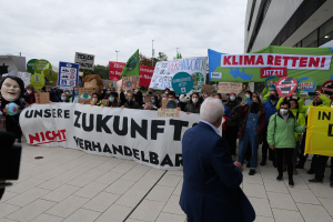 Eine Gruppe von Menschen hält Schilder und Transparente und protestiert gegen den Klimawandel vor einem Gebäude in Deutschland, mit einer Person, die links eine Kamera hält, Bäumen, Pfählen und einem klaren blauen Himmel im Hintergrund.