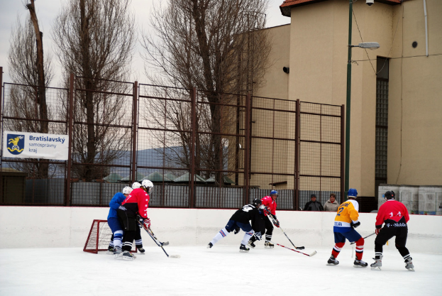 Personen beim Eisschnelllauf auf einer Eisfläche mit Gebäuden, Bäumen, einer Straßenlaterne, einem Namensschild und Zäunen im Hintergrund unter dem Himmel.