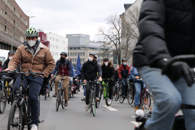 Eine Gruppe von Menschen in Helmen und Handschuhen fährt auf Fahrrädern eine von Bäumen gesäumte Straße in Berlin, Deutschland, entlang, wobei Gebäude und ein geparktes Fahrzeug im Hintergrund zu sehen sind.