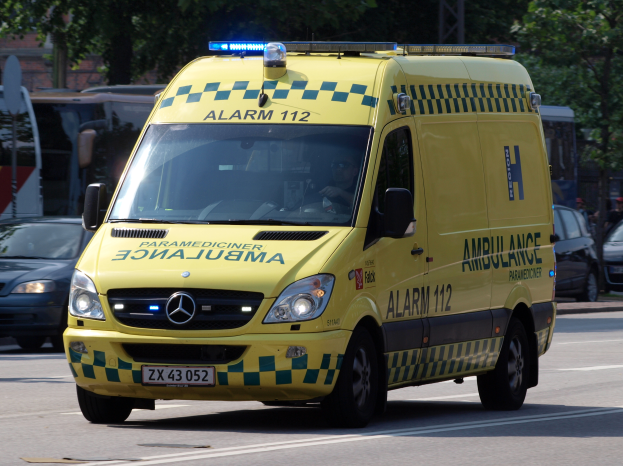 Ein Rettungswagen mit eingeschaltetem Blaulicht fährt auf einer Straße, umgeben von anderen Fahrzeugen, mit Bäumen, Pfählen und einem Gebäude im Hintergrund.