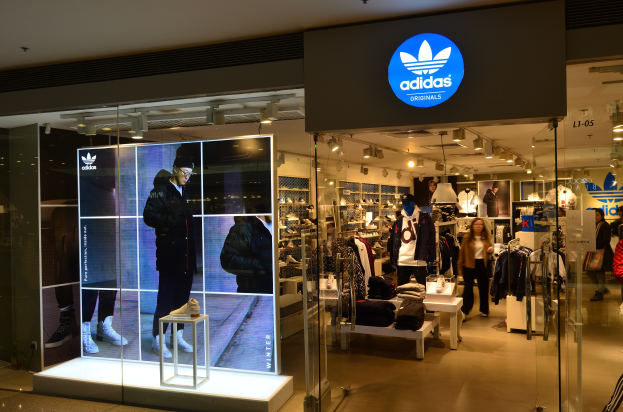 Ein Adidas-Laden in einem Einkaufszentrum mit Schaufensterpuppen, Kleider an Bügeln, Schuhe auf Ständern und beleuchteter Laden mit einem Schild mit dem Adidas-Logo, das auf die Adidas Originals-Marke hinweist.
