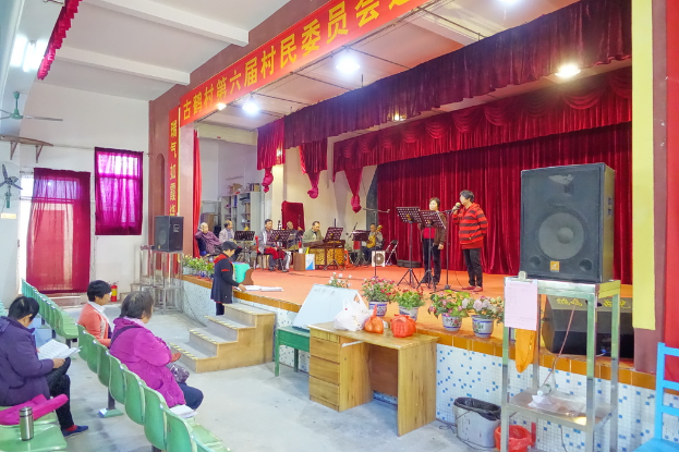 Eine Bühne mit zwei singenden Personen, einer Band im Hintergrund, roten Vorhängen, einem Scheinwerfer, einem Lautsprecher auf der rechten Seite und einem sitzenden Publikum.