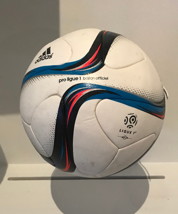 Ein weißer Füßball mit dem Adidas-Logo und "Ligue 1" in fetter schwarzer Schrift auf einem weißen Tisch gegen einen hellweißen Hintergrund.