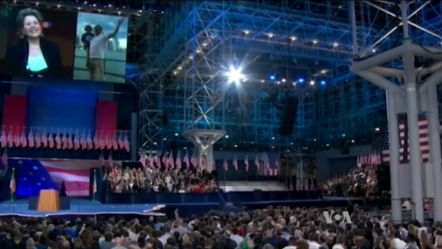 Eine große Menschenmenge sitzt vor einer Bühne mit einem Podium, einem Mikrofon, Flaggen, einem Bildschirm und Beleuchtungsausrüstung und wartet auf eine Präsidentschaftsdiskussion.