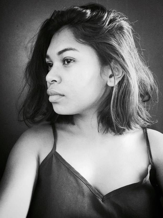 Eine selbstbewusste Frau in einem schwarzen Kleid posiert in einem Schwarz-Weiß-Foto, ihr glatter Bob und das natürliche Make-up unterstreichen ihre selbstbewusste Haltung vor einer Wand.