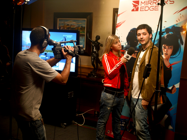 Ein Mann mit einer Videokamera und einem Headset filmt ein Interview zwischen einem Mann und einer Frau, mit einem Fernseher und einem Rahmen im Hintergrund.