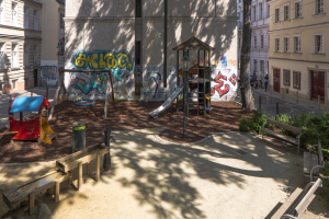 Ein Spielplatz mit Graffiti in einer städtischen Umgebung, umgeben von Gebäuden, Bäumen, Bänken und einem Mülleimer, mit Menschen und Fahrzeugen auf der Straße und dem Himmel im Hintergrund.