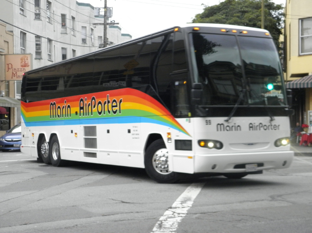 Ein Bus fährt auf einer Straße mit Gebäuden, Bäumen und Mästen im Hintergrund.