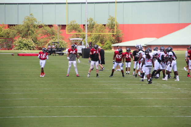 Eine Gruppe von Menschen, die Fußball auf einem Feld spielen, das von Bäumen, Pfählen, Hütten und einem klaren blauen Himmel umgeben ist, wobei einige Spieler Helme tragen.
