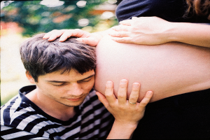 Ein Mann und eine Frau stehen nebeneinander, der Mann berührt sanft den Bauch der Frau, mit einem unscharfen Hintergrund, der das Paar und ihr ungeborenes Kind hervorhebt.