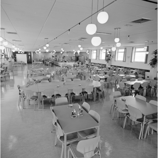 Eine geräumige Kantine mit Reihen von Tischen und Stühlen, verstreute Flaschen und Gegenstände auf den Tischen, Deckenbeleuchtung, Fenster an der rechten Wand und Topfpflanzen überall im Raum.