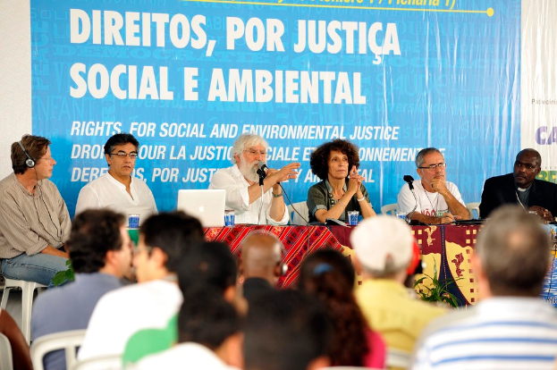 Sechs Personen sitzen an einem Tisch auf einer Bühne mit Gläsern, einem Laptop und Mikrofonen, gegenüber einem Publikum mit einem Banner im Hintergrund.