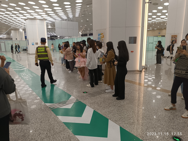 Eine Gruppe von Menschen steht in einer Schlange an einem Flughafen-Check-in, einige halten Taschen und Mobiltelefone, mit Säulen im Hintergrund und Lampen an der Decke und einem Wasserzeichen am unteren Rand des Bildes.