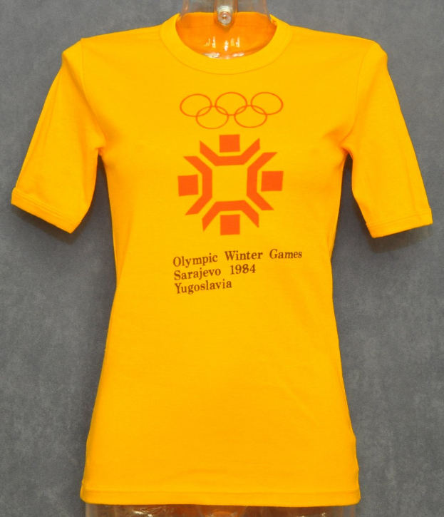 Ein gelbes T-Shirt mit der Aufschrift "Olympic Winter Games 1984", das an einer Schaufensterpuppe an einer Wand hängt.