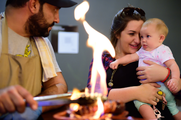 Ein Mann mit schwarzer Mütze kocht Essen über einem Feuer, während eine strahlende Frau ein Baby in einem weißen Oberteil und grünen Hosen hält.