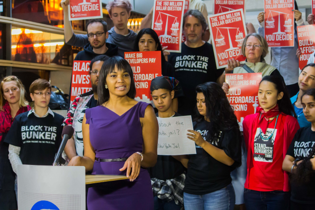 Eine Black-Lives-Matter-Aktivistin spricht vor einem Publikum mit Mikrofon an einem Rednerpult, das Publikum hält Schilder, es gibt Lichter und andere Gegenstände im Hintergrund.