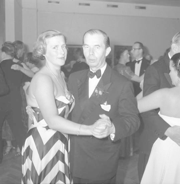 Ein Mann in einem schwarzen Blazer, weißem Hemd und schwarzen Hosen tanzt mit einer Frau in unbekannter Kleidung auf einer formellen Veranstaltung, indem sie sich an den Händen halten und zur Musik an einer Wand tanzen.