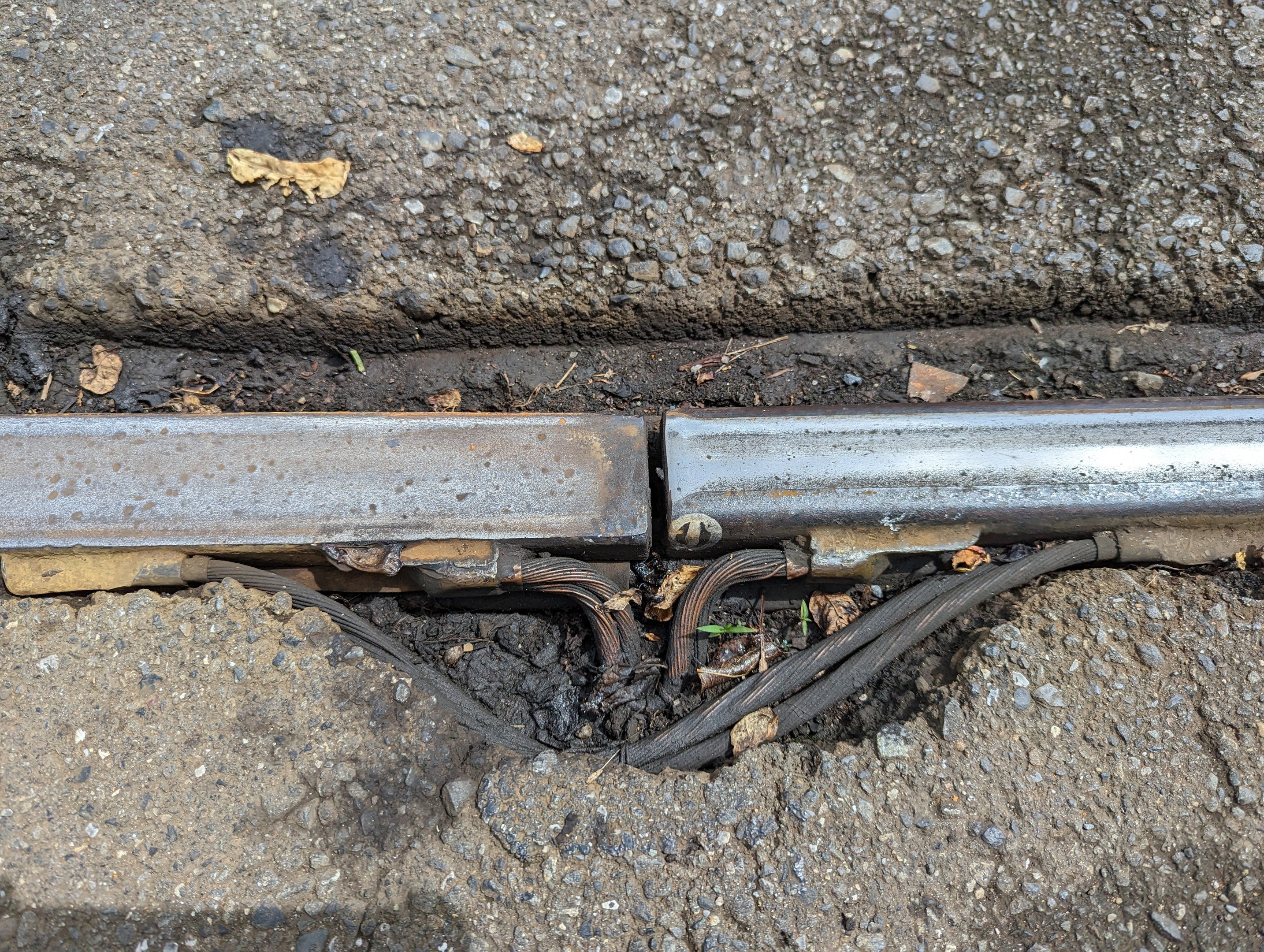 Nahaufnahme eines Metallkanalrohrs auf der Straße mit daran befestigten Drähten und verstreuten trockenen Blättern.