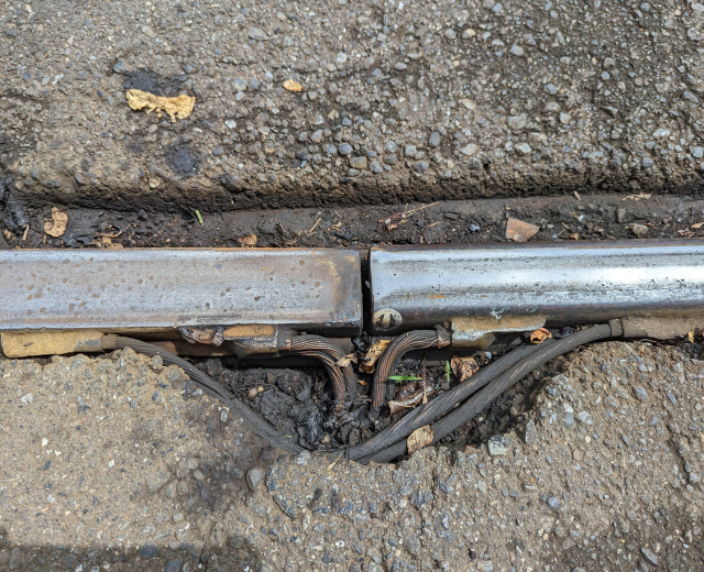Nahaufnahme eines Metallkanalrohrs auf der Straße mit daran befestigten Drähten und verstreuten trockenen Blättern.