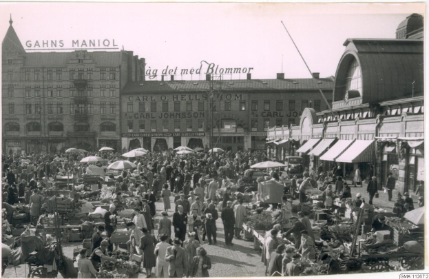 Altes Schwarz-Weiß-Foto eines belebten Berliner Markts mit Menschen, Gemüsewagen, Schirmen und Gebäuden mit Fenstern im Hintergrund.
