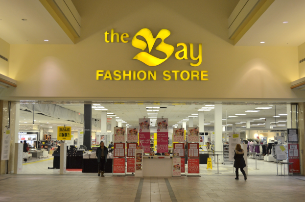 Außenansicht eines Bay Fashion Stores in einem Einkaufszentrum mit Menschen in der Nähe, Texttafeln an Wänden, Säulen, Deckenleuchten und Hintergrundanzeigen.