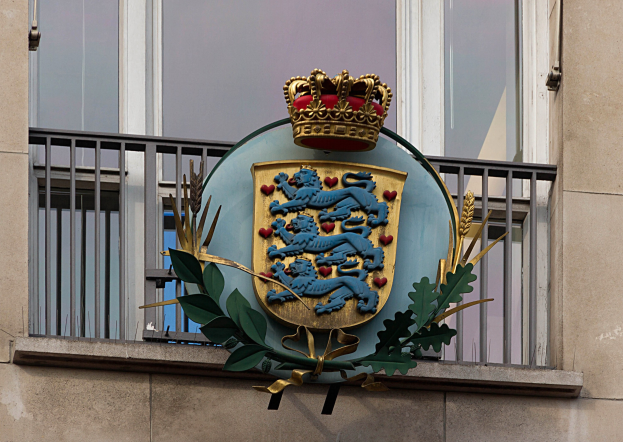 Das britische Königswappen auf der Außenseite eines Gebäudes, begleitet von einem Logo mit Krone und Blättern, mit sichtbaren Fenstern und Gittern im Hintergrund.