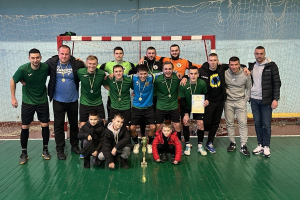 Eine Gruppe von Männern in Medaillen steht zusammen auf einem Futsal-Platz, einer hält einen Pokal, mit einem Torpfosten und einer Wand im Hintergrund.