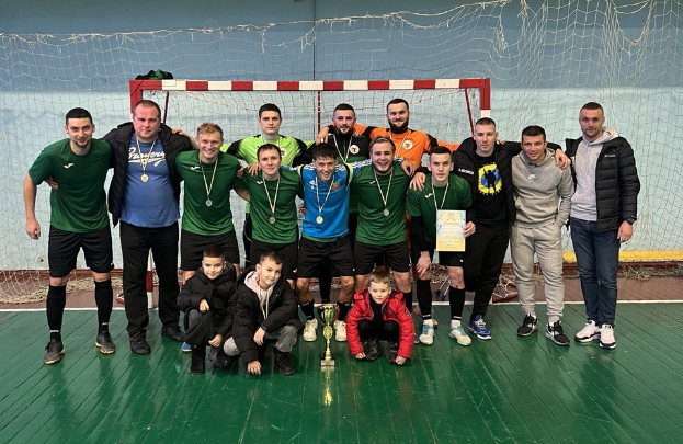 Eine Gruppe von Männern in Medaillen steht zusammen auf einem Futsal-Platz, einer hält einen Pokal, mit einem Torpfosten und einer Wand im Hintergrund.