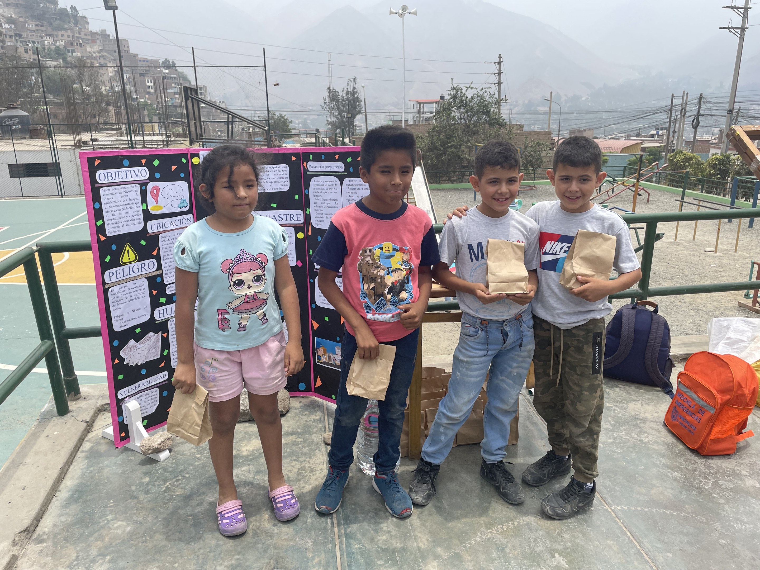 Gruppe von Kindern auf einem Basketballplatz mit Papiertüten in den H├Ąnden, mit Tüten und Gegenst├Ąnden auf der rechten Seite und einer Tafel mit Text auf der linken Seite, vor einem Hintergrund aus Z├Ąunen, Pfosten, Stra├čenlaternen, B├Ąumen, Geb├Ąuden, H├Ąngen und Himmel.