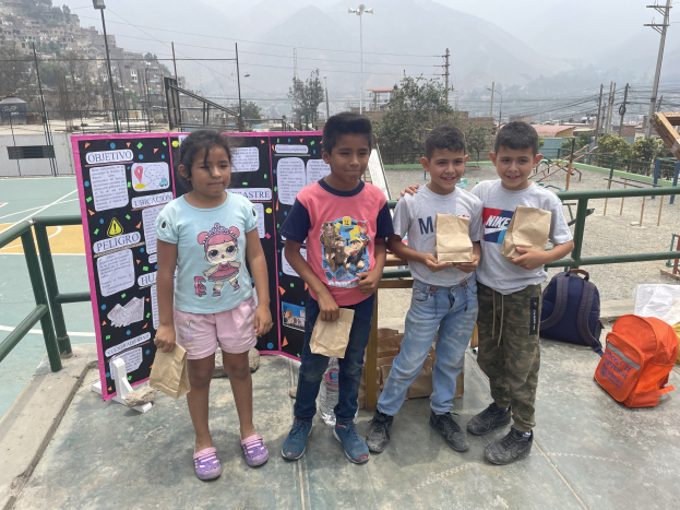Gruppe von Kindern auf einem Basketballplatz mit Papiertüten in den H├Ąnden, mit Tüten und Gegenst├Ąnden auf der rechten Seite und einer Tafel mit Text auf der linken Seite, vor einem Hintergrund aus Z├Ąunen, Pfosten, Stra├čenlaternen, B├Ąumen, Geb├Ąuden, H├Ąngen und Himmel.