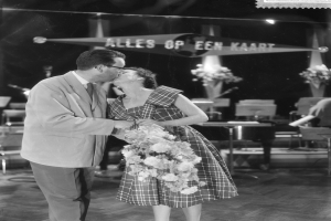 Ein Mann und eine Frau umarmen und küssen sich auf einer Bühne, wobei die Frau einen Blumenstrauß hält, während Stühle und Blumenvasen im Hintergrund zu sehen sind.