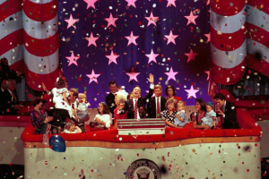 Eine Gruppe von Menschen steht auf einer Bühne mit einem Rednerpult, Ballons und Konfetti, mit Sternen und einem Vorhang im Hintergrund, was auf eine Präsidentenamtseinführung hindeutet.