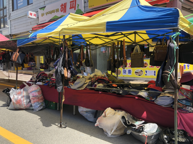 Ein Straßenmarkt mit Zelten, die Taschen, Hüte und andere Gegenstände präsentieren, sowie Gebäuden mit Fenstern und Texttafeln.