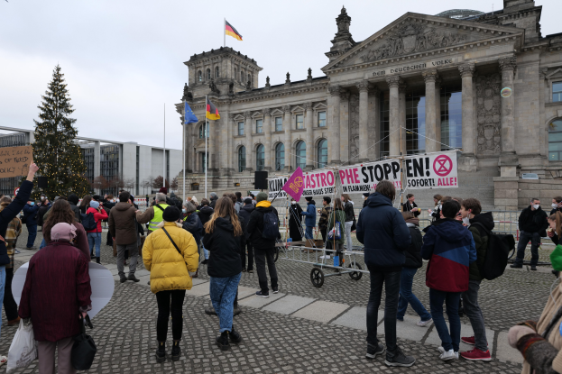 Eine Gruppe von Menschen steht vor einem Gebäude mit Fenstern, Säulen und Bögen und hält Schilder und Fahnen, wahrscheinlich protestierend in Reaktion auf den Reichstag, mit einem Baum im Vordergrund und einem bewölkten Himmel im Hintergrund.